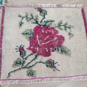 Puede incluir: Una peque&ntilde;a alfombra rectangular con fondo crema y un gran dise&ntilde;o de rosa magenta. Hojas y tallos verdes rodean la flor. La alfombra tiene un borde rosa y flecos.
