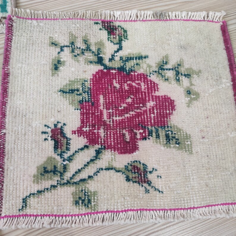 Puede incluir: Una peque&ntilde;a alfombra rectangular con fondo crema y un gran dise&ntilde;o de rosa magenta. Hojas y tallos verdes rodean la flor. La alfombra tiene un borde rosa y flecos.