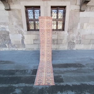 Könnte beinhalten: Ein langer, schmaler Teppich mit einem farbenfrohen geometrischen Muster in Rot-, Orange-, Gelb- und Brauntönen. Der Teppich liegt auf einem grauen Betonboden vor einer Steinwand mit zwei Fenstern.