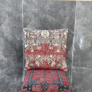 May include: A decorative pillow with a red and beige patterned design, resting on a matching cushion. The pillow features a detailed, vintage-style pattern with a central red field and ornate borders. The cushion below has a similar design with teal accents.