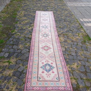 Peut inclure: Un long tapis rectangulaire avec un champ central cr&egrave;me et un motif g&eacute;om&eacute;trique r&eacute;p&eacute;titif dans des tons de rose, de bleu et de gris. Le tapis a une bordure rose et est expos&eacute; &agrave; l'ext&eacute;rieur sur une surface pav&eacute;e.