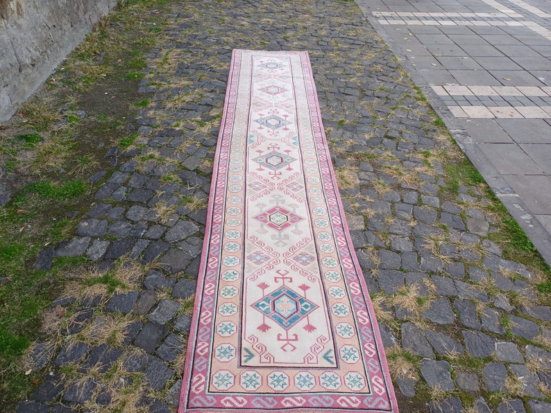 Peut inclure: Un long tapis rectangulaire avec un champ central cr&egrave;me et un motif g&eacute;om&eacute;trique r&eacute;p&eacute;titif dans des tons de rose, de bleu et de gris. Le tapis a une bordure rose et est expos&eacute; &agrave; l'ext&eacute;rieur sur une surface pav&eacute;e.