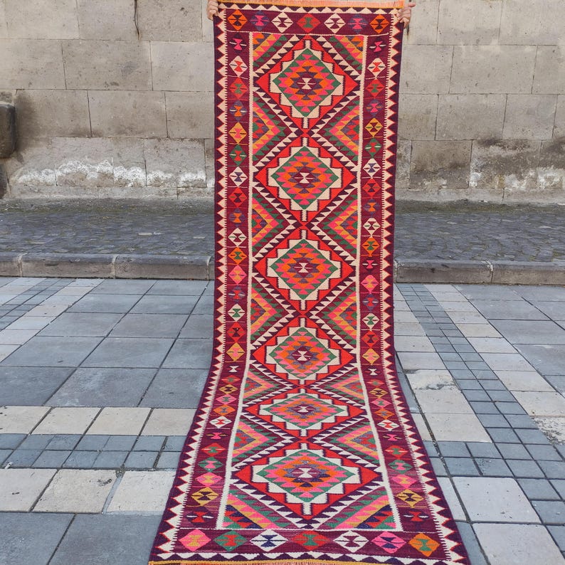 May include: A long, rectangular rug with a geometric pattern. The rug features a deep burgundy background with a repeating pattern of diamonds in shades of orange, green, and pink. The edges are trimmed with a zig-zag design.