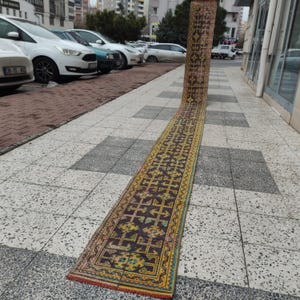 Może przedstawiać: Długi, wąski, prostokątny dywan z geometrycznym wzorem w odcieniach brązu, żółci i turkusu. Dywan jest zwinięty na jednym końcu i leży na kafelkowej powierzchni, prezentując swój skomplikowany wzór i żywe kolory.
