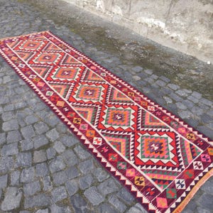May include: A long, rectangular rug with a geometric pattern. The rug features a repeating design of red, orange, green, and pink diamond shapes on a burgundy background. The edges are finished with a white and red zigzag pattern.