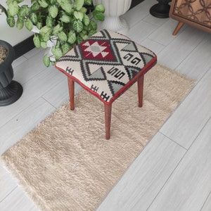 Può includere: Un pouf quadrato con un top in tessuto a motivi geometrici in tonalità di rosso, grigio e bianco. Il pouf ha quattro gambe in legno.
