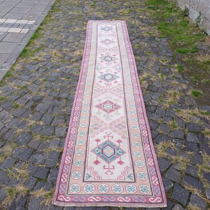 Peut inclure: Un long tapis rectangulaire avec un champ central cr&egrave;me et une bordure rose. Le tapis pr&eacute;sente des motifs g&eacute;om&eacute;triques dans des tons de rose, turquoise et gris. Le tapis est pos&eacute; sur une surface pav&eacute;e.