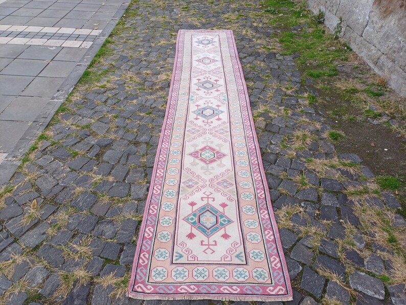 Peut inclure: Un long tapis rectangulaire avec un champ central cr&egrave;me et une bordure rose. Le tapis pr&eacute;sente des motifs g&eacute;om&eacute;triques dans des tons de rose, turquoise et gris. Le tapis est pos&eacute; sur une surface pav&eacute;e.