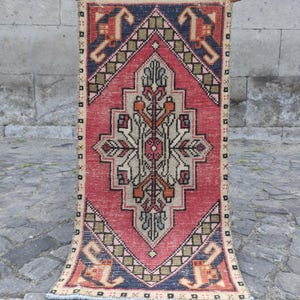 May include: A rectangular woven rug featuring a large central diamond motif in cream, surrounded by a vibrant red field. The design includes geometric patterns in navy blue, olive green, and orange, with a decorative border of small squares.