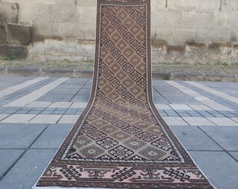 May include: A long, narrow rug with a geometric pattern in shades of brown, pink, and black. The rug has a fringe border and is laid out on a gray tiled floor.