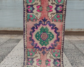 May include: A rectangular woven rug featuring a central geometric medallion in green, blue, and pink. The background is a faded salmon pink with symmetrical floral-inspired motifs in cream, teal, and dark blue, finished with a dark blue border.