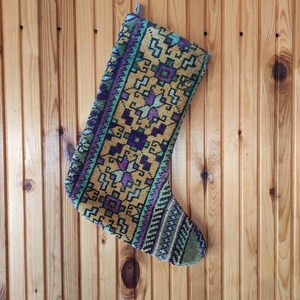 Può includere: Una calza di Natale con un motivo geometrico nei toni dell'oro, del viola, del verde e del verde acqua. La calza è appesa su uno sfondo di legno. Il design presenta forme e linee ripetute.