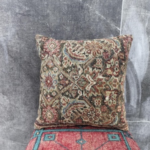 Op de afbeelding: Vierkant kussen met een vintage tapijtontwerp. Het kussen heeft een complex patroon in tinten bruin, rood, groen en blauw. Het kussen ligt op een rood tapijt met een turquoise rand.