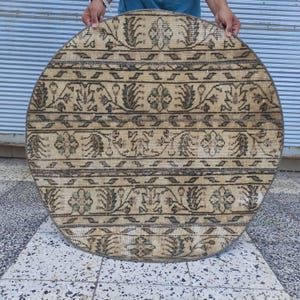 May include: A circular rug featuring a beige background with repeating horizontal bands of dark brown and muted green floral motifs. The design consists of alternating rows of intricate botanical patterns and geometric lines across the surface.