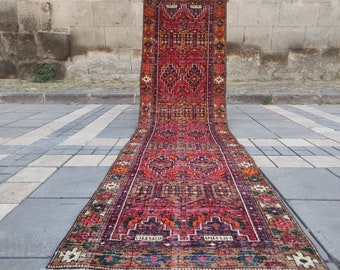 May include: A long, narrow, red, blue and orange patterned rug with a geometric design. The rug is laid out on a grey pavement.