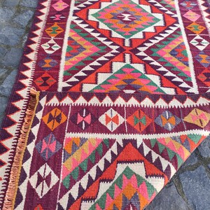 May include: A colorful, geometric patterned rug with a folded corner. The rug features a variety of shapes and colors, including red, pink, green, orange, and white. The rug has a fringed edge and is likely made of woven material.