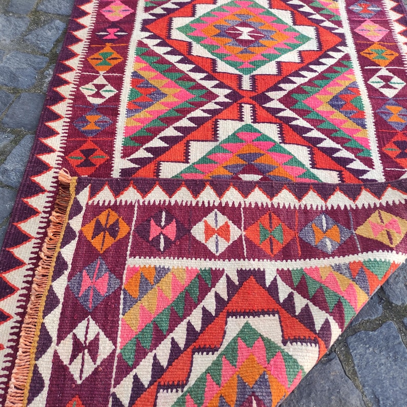 May include: A colorful, geometric patterned rug with a folded corner. The rug features a variety of shapes and colors, including red, pink, green, orange, and white. The rug has a fringed edge and is likely made of woven material.