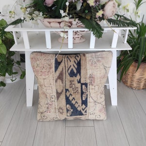 Può includere: Un cuscino decorativo in tessuto beige e marrone con motivi. Il cuscino ha una striscia blu scuro al centro ed è appoggiato su uno scaffale bianco.