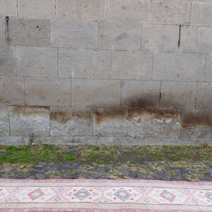 Peut inclure: Un tapis rectangulaire avec une bordure rose et un champ central cr&egrave;me. Le tapis pr&eacute;sente un motif g&eacute;om&eacute;trique r&eacute;p&eacute;titif en rose, bleu et cr&egrave;me. Le tapis est pos&eacute; sur une surface pav&eacute;e devant un mur de pierre grise.
