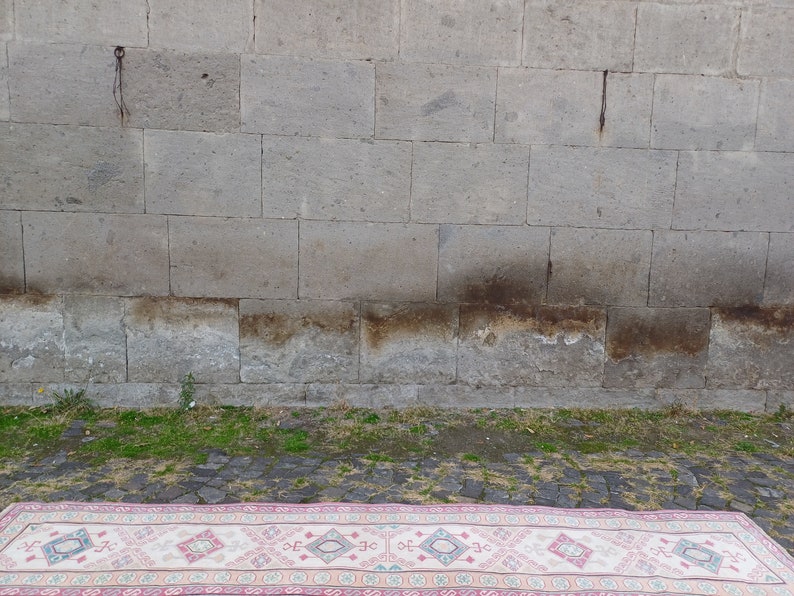 Peut inclure: Un tapis rectangulaire avec une bordure rose et un champ central cr&egrave;me. Le tapis pr&eacute;sente un motif g&eacute;om&eacute;trique r&eacute;p&eacute;titif en rose, bleu et cr&egrave;me. Le tapis est pos&eacute; sur une surface pav&eacute;e devant un mur de pierre grise.