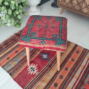 Könnte beinhalten: Ein roter, blauer und grüner gemusterter Hocker mit Holzbeinen steht auf einem bunten Teppich mit geometrischen Mustern.