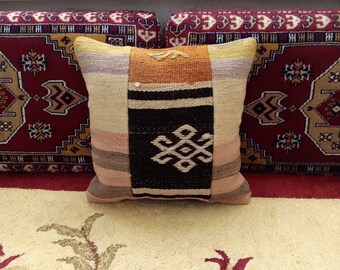 May include: A colorful woven pillow with a geometric pattern in shades of beige, brown, orange, and pink. The pillow is sitting on a red and beige patterned rug.