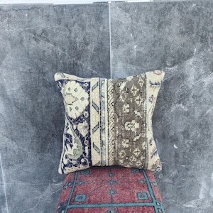 May include: A decorative square pillow with a patchwork design. The pillow features a combination of beige, navy blue, and brown patterns. The pillow is placed on top of a red and blue patterned surface.