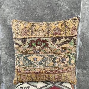 Op de afbeelding: Een vierkant decoratief kussen met een vintage tapijtontwerp. Het kussen heeft een mix van kleuren, waaronder goud, bruin, groen en rood. Het patroon bevat bloemen en geometrische elementen, waardoor een unieke en getextureerde uitstraling ontstaat. Het kussen wordt waarschijnlijk gebruikt voor woondecoratie.