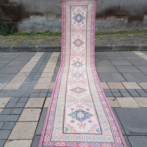 Peut inclure: Un long tapis de couloir rectangulaire avec un champ central cr&egrave;me et un motif g&eacute;om&eacute;trique r&eacute;p&eacute;titif en rose, bleu et turquoise. Le tapis a une bordure rose et est expos&eacute; &agrave; l'ext&eacute;rieur sur une surface en pierre.