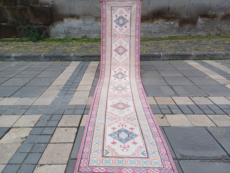 Peut inclure: Un long tapis de couloir rectangulaire avec un champ central cr&egrave;me et un motif g&eacute;om&eacute;trique r&eacute;p&eacute;titif en rose, bleu et turquoise. Le tapis a une bordure rose et est expos&eacute; &agrave; l'ext&eacute;rieur sur une surface en pierre.