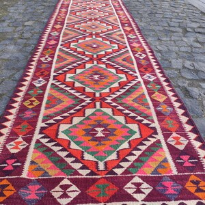 May include: A long, rectangular runner rug with a geometric pattern. The rug features a burgundy border and a central design with diamonds in shades of red, orange, green, and white. The rug is likely made of woven fabric.