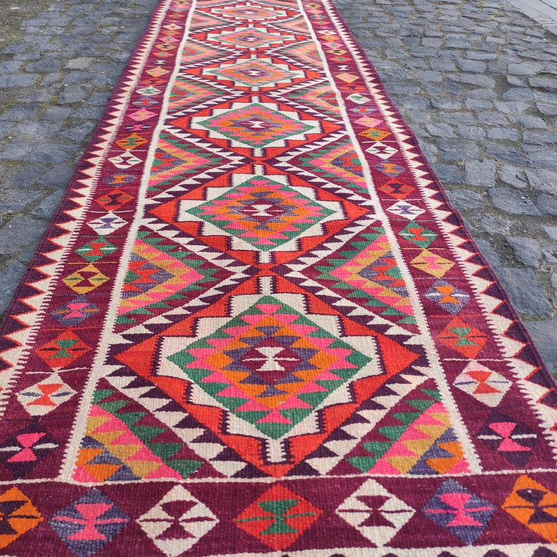 May include: A long, rectangular runner rug with a geometric pattern. The rug features a burgundy border and a central design with diamonds in shades of red, orange, green, and white. The rug is likely made of woven fabric.