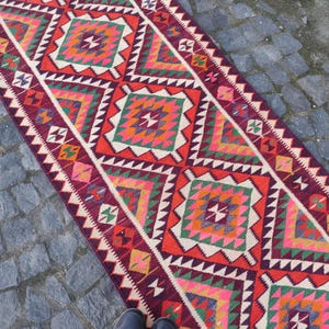 May include: A vibrant, rectangular rug with a geometric pattern. The rug features a repeating design of diamond shapes in red, orange, green, pink, and white. The edges are bordered with a zigzag pattern. The rug is placed on a stone surface.