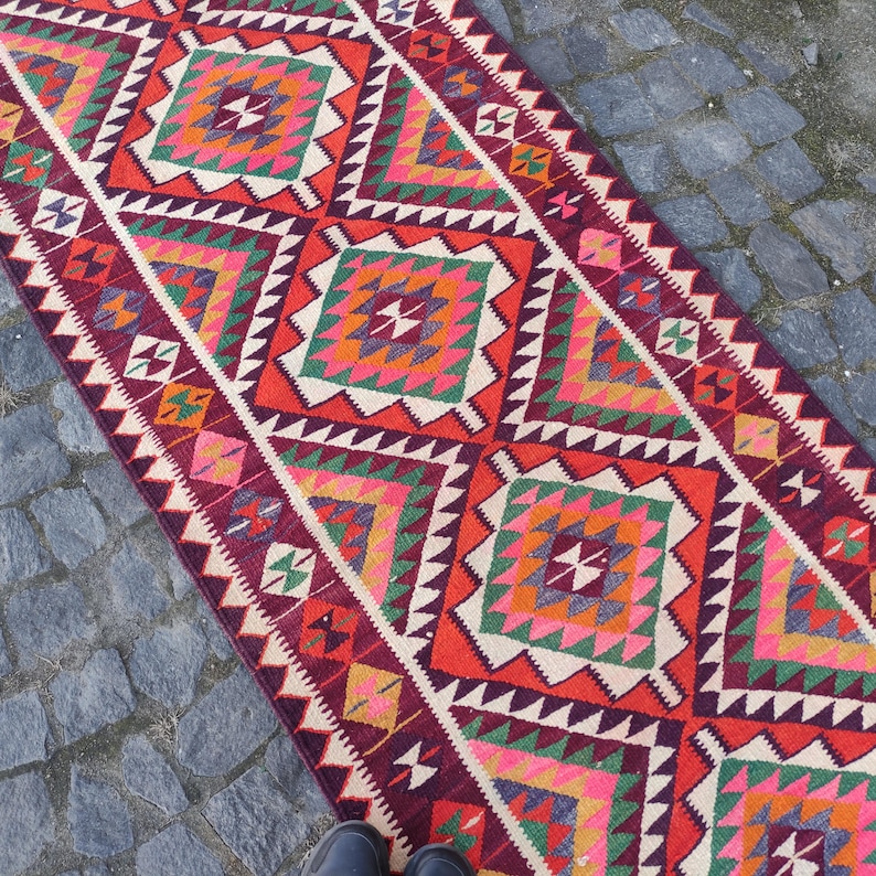 May include: A vibrant, rectangular rug with a geometric pattern. The rug features a repeating design of diamond shapes in red, orange, green, pink, and white. The edges are bordered with a zigzag pattern. The rug is placed on a stone surface.
