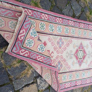 Peut inclure: Un tapis rectangulaire pli&eacute;, avec un champ central cr&egrave;me et une bordure rose. Le tapis pr&eacute;sente des motifs g&eacute;om&eacute;triques roses, bleus et p&ecirc;che. La frange du tapis est visible dans le coin sup&eacute;rieur gauche. Le tapis est pos&eacute; sur une surface en pierre.