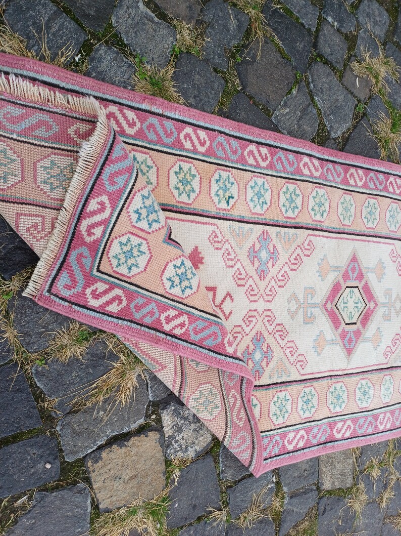 Peut inclure: Un tapis rectangulaire pli&eacute;, avec un champ central cr&egrave;me et une bordure rose. Le tapis pr&eacute;sente des motifs g&eacute;om&eacute;triques roses, bleus et p&ecirc;che. La frange du tapis est visible dans le coin sup&eacute;rieur gauche. Le tapis est pos&eacute; sur une surface en pierre.