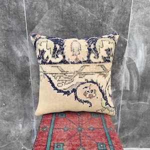 Può includere: Cuscino quadrato decorativo con design a tappeto vintage. Il cuscino presenta un centro color crema con un bordo blu navy e accenti verdi e marroni. È posto su un tappeto rosso con un motivo geometrico.