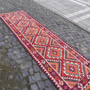 May include: A long, rectangular runner rug with a geometric pattern. The rug features a repeating design of diamonds and triangles in shades of red, orange, green, and white. The rug is placed on a cobblestone surface.