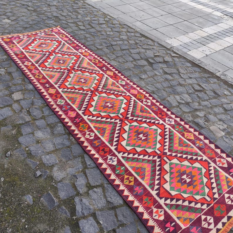 May include: A long, rectangular runner rug with a geometric pattern. The rug features a repeating design of diamonds and triangles in shades of red, orange, green, and white. The rug is placed on a cobblestone surface.