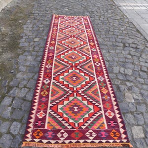 May include: A long, rectangular rug with a geometric pattern. The rug features a central design of diamonds and triangles in shades of red, orange, green, and white. The edges are a deep burgundy with a fringe.