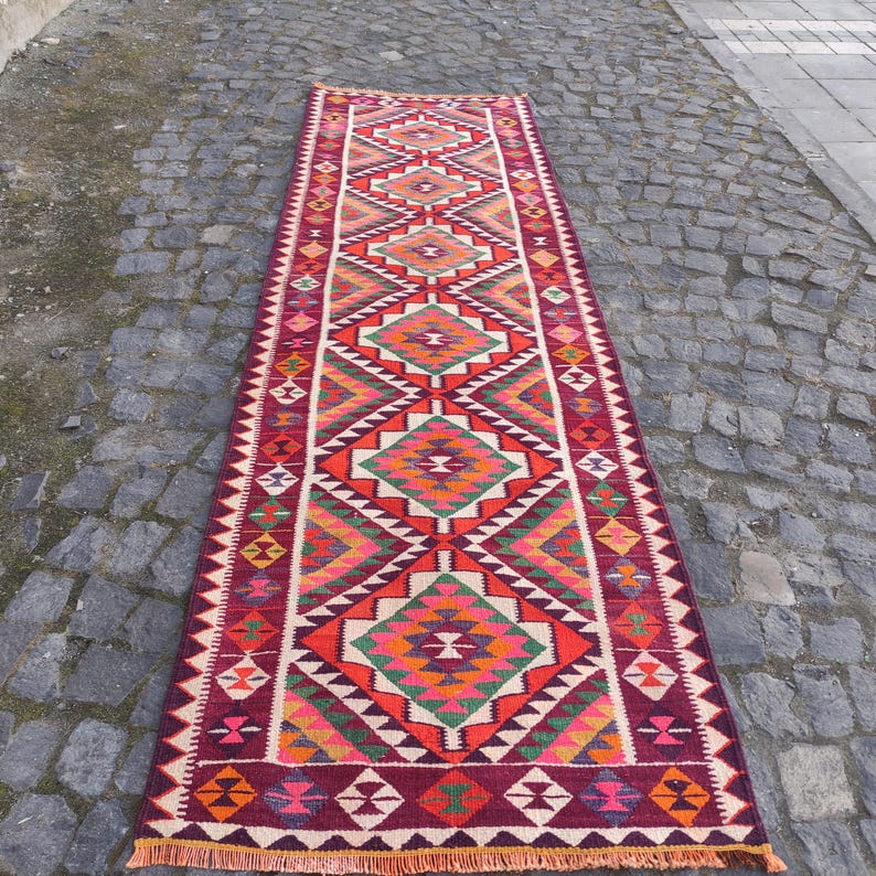 May include: A long, rectangular rug with a geometric pattern. The rug features a central design of diamonds and triangles in shades of red, orange, green, and white. The edges are a deep burgundy with a fringe.