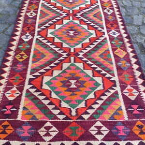 May include: A rectangular, hand-woven rug with a geometric pattern. The rug features a central diamond design in shades of orange, green, pink, and purple, framed by white and burgundy borders. The rug's design is symmetrical and detailed.