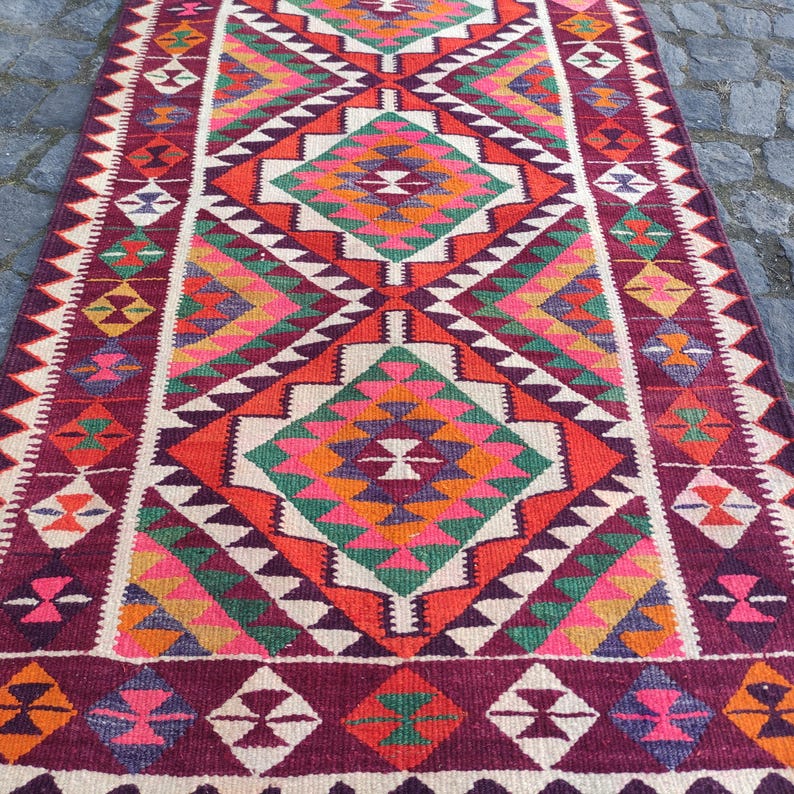 May include: A rectangular, hand-woven rug with a geometric pattern. The rug features a central diamond design in shades of orange, green, pink, and purple, framed by white and burgundy borders. The rug's design is symmetrical and detailed.