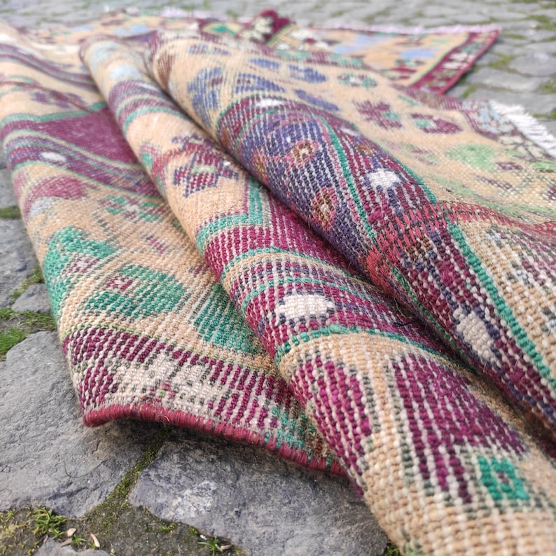 May include: A vintage rug with a geometric pattern in shades of beige, green, red, and purple. The rug is folded in half, showing the intricate details of the woven design.