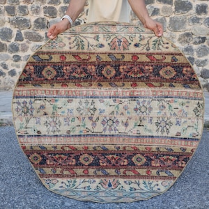 May include: A round rug featuring a patchwork design with horizontal bands of varying patterns. The rug displays a color palette of beige, dark brown, red, blue, and green, with intricate floral and geometric motifs throughout the woven surface.