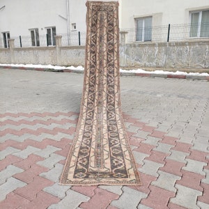 May include: A long, narrow rug with a beige background and brown geometric patterns. The rug is folded in half and lying on a gray brick surface.