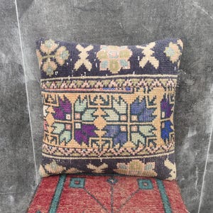 May include: A square, decorative pillow with a dark blue background and a central geometric pattern in beige, purple, and green. The pillow is placed on a red patterned surface, creating a contrast in colors and textures.
