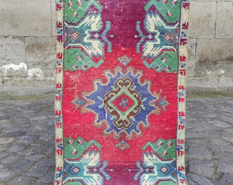 May include: A rectangular woven rug featuring a vibrant red and magenta field with a central blue geometric medallion. The design includes green and teal stylized floral motifs at the corners and a cream-colored border with small geometric patterns.