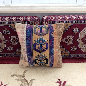 May include: A decorative pillow with a colorful geometric pattern in shades of blue, yellow, pink, and purple. The pillow is made of a woven fabric with a distressed look. The pillow is sitting on a rug with a similar pattern.