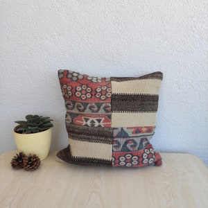 Può includere: Cuscino decorativo quadrato con design patchwork. Il cuscino presenta un mix di motivi e colori, tra cui rosso, marrone, beige e grigio. Il cuscino è posto su una superficie di legno chiaro.